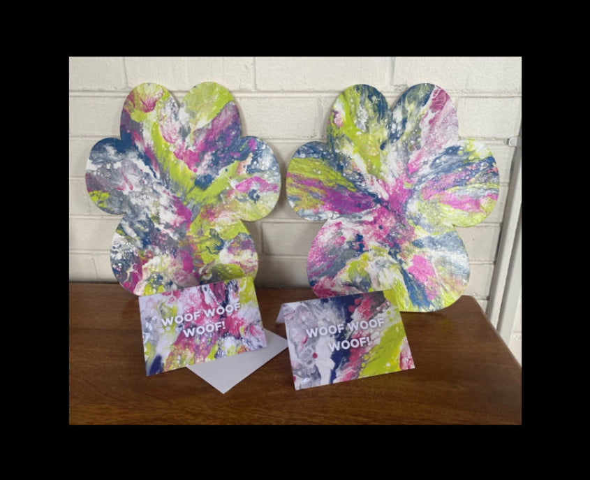 Two large paw-shaped art prints with a colorful abstract paint splatter design, displayed on a wooden table in front of a white brick wall. Each print includes a small matching card underneath with the text 'WOOF WOOF WOOF!'.